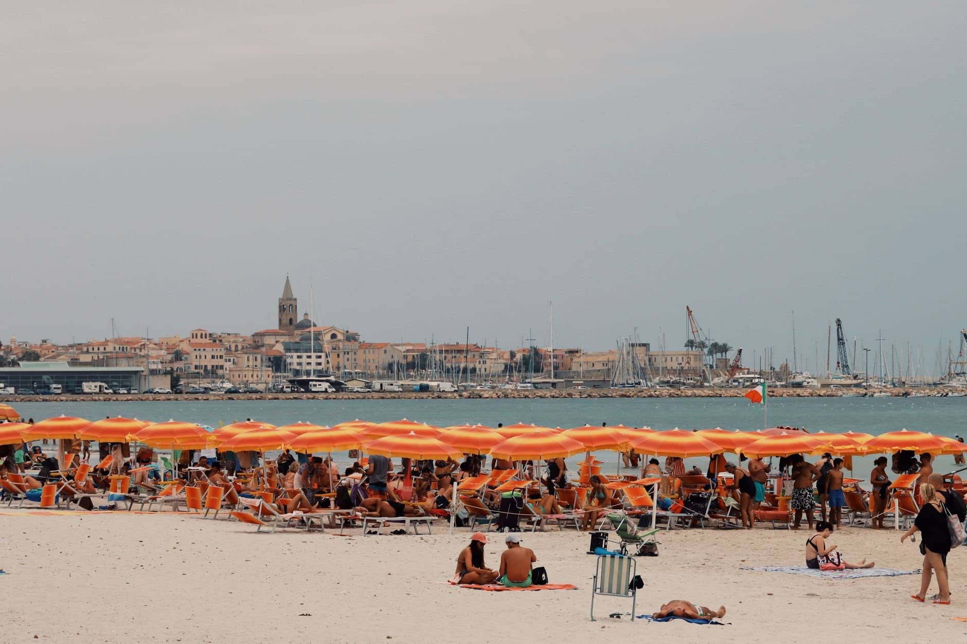 Crowded Alghero beach with orange umbrellas and sea view in Sardinia, Italy.
