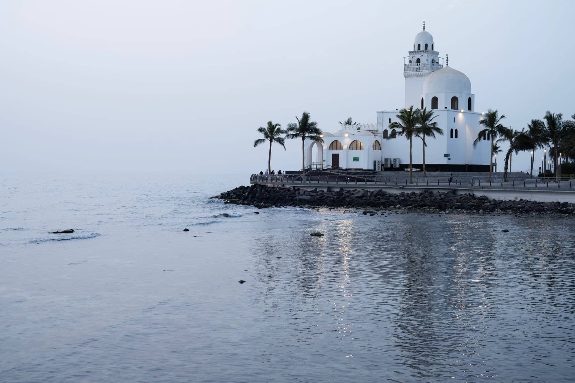 Scenic view of a seaside mosque in Jeddah, Saudi Arabia with palm trees and calm waters.