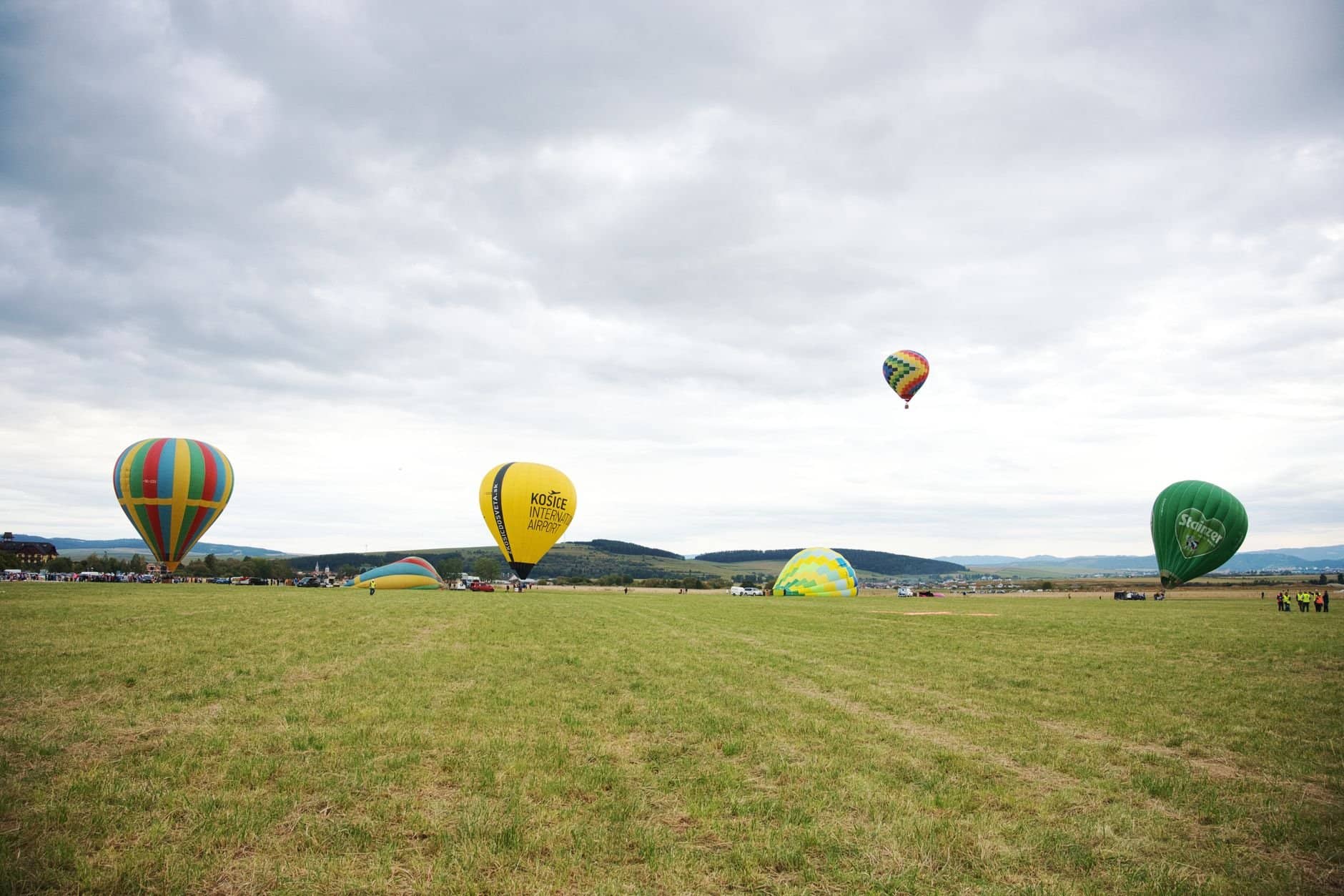 Hot air balloons soar over a scenic grass field in Poprad, Slovakia, under a clouded sky.