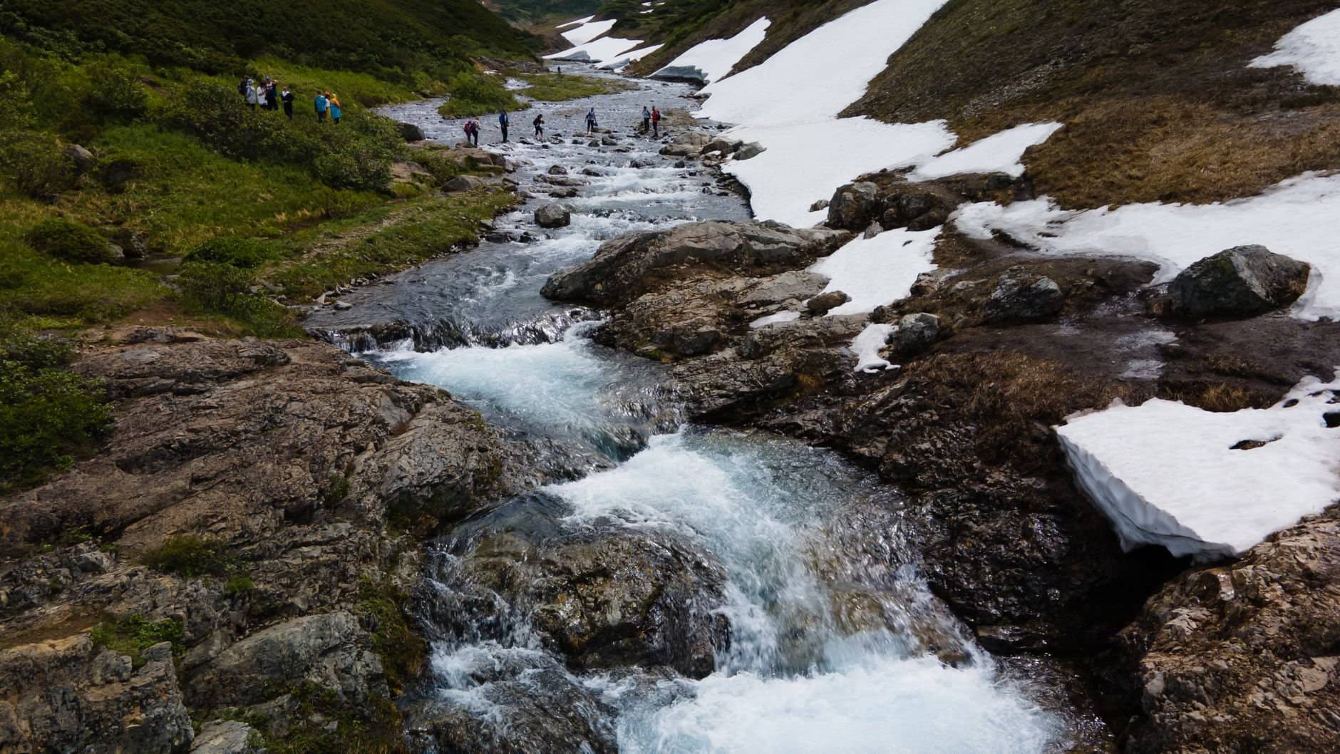 A breathtaking view of a flowing mountain stream surrounded by snow patches and hikers in Kamchatka.