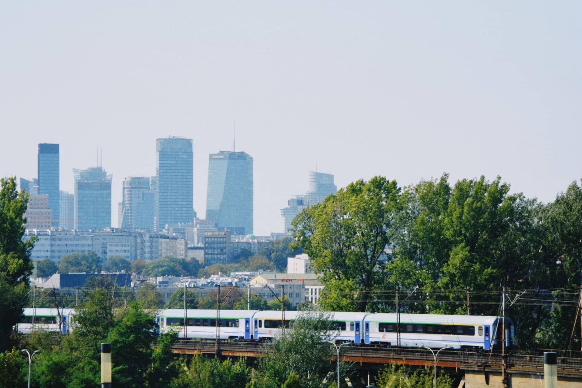 Skyline of Warsaw with modern buildings and a train passing amidst the greenery.