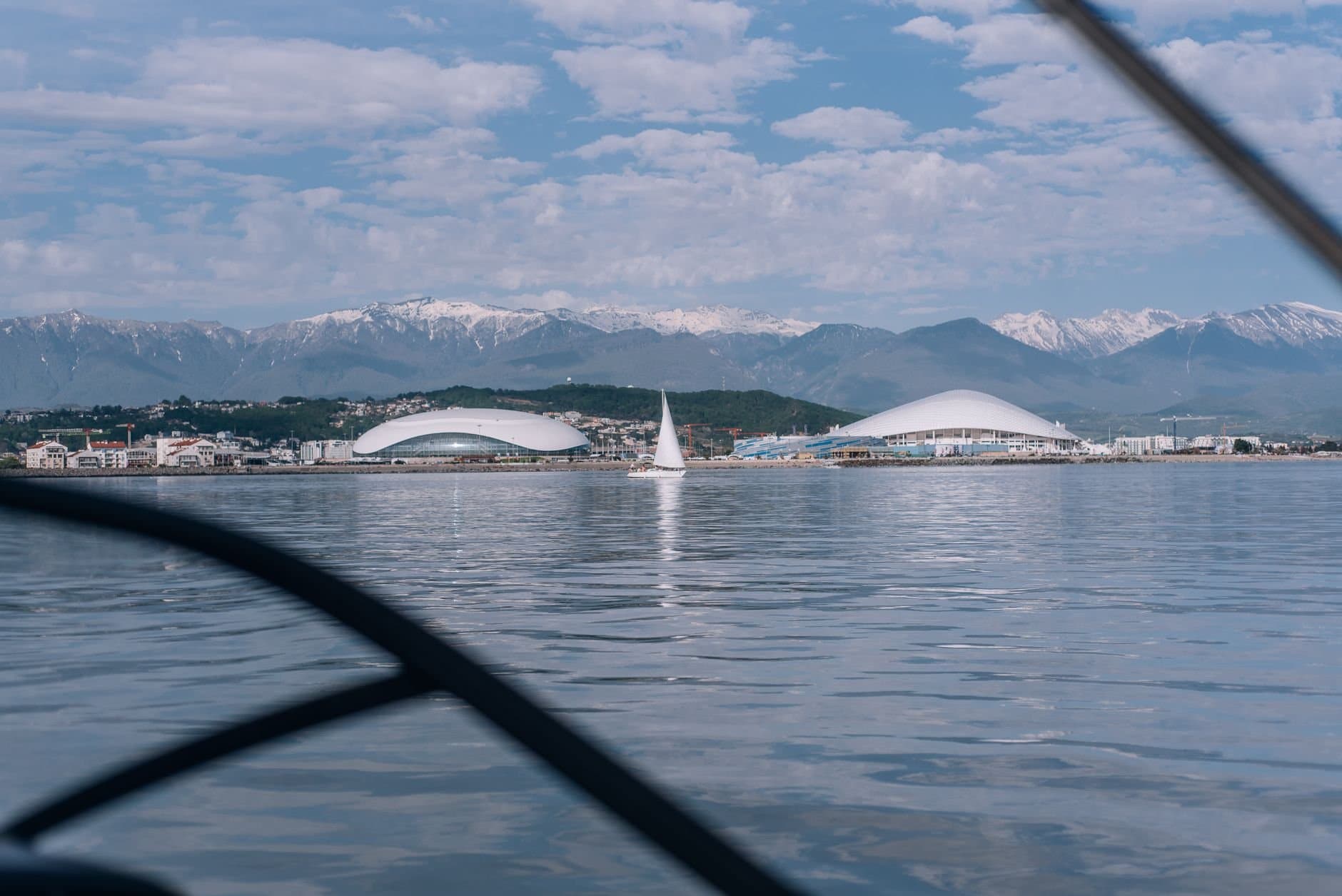 A breathtaking view of the Sochi Olympic Park with snow-capped mountains and a sailboat on the water.