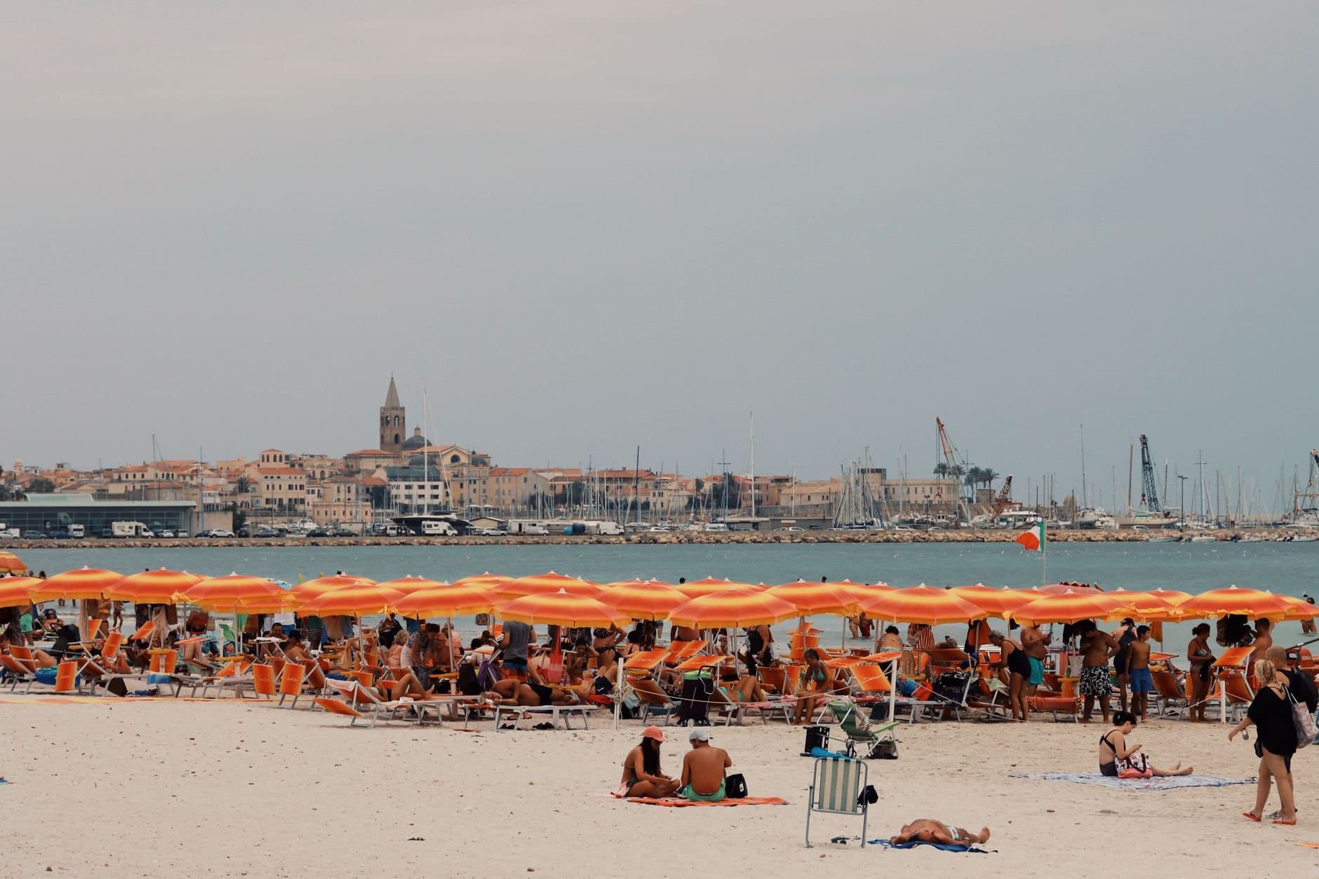 Crowded Alghero beach with orange umbrellas and sea view in Sardinia, Italy.