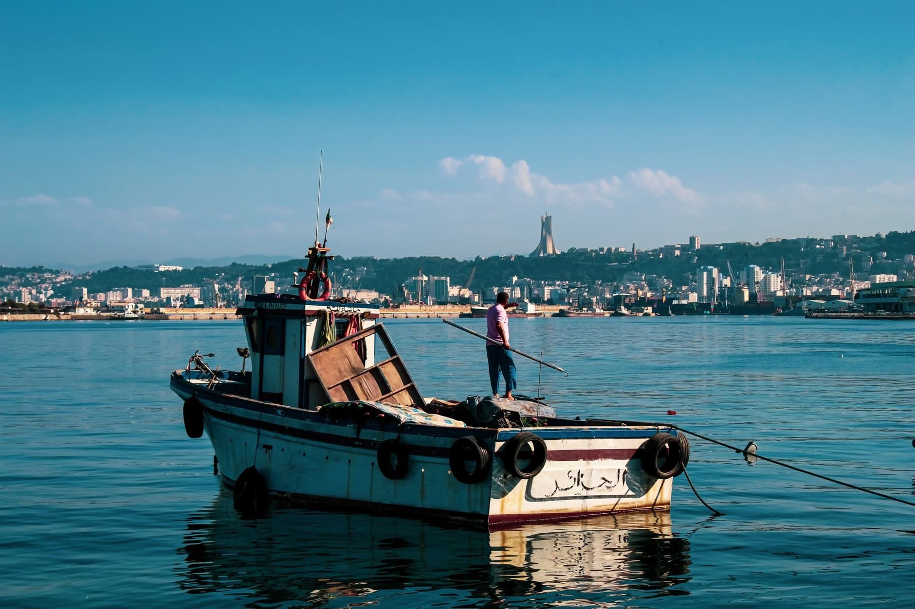 A fishing boat in Algiers Harbor with the city's skyline, including a fisherman onboard.