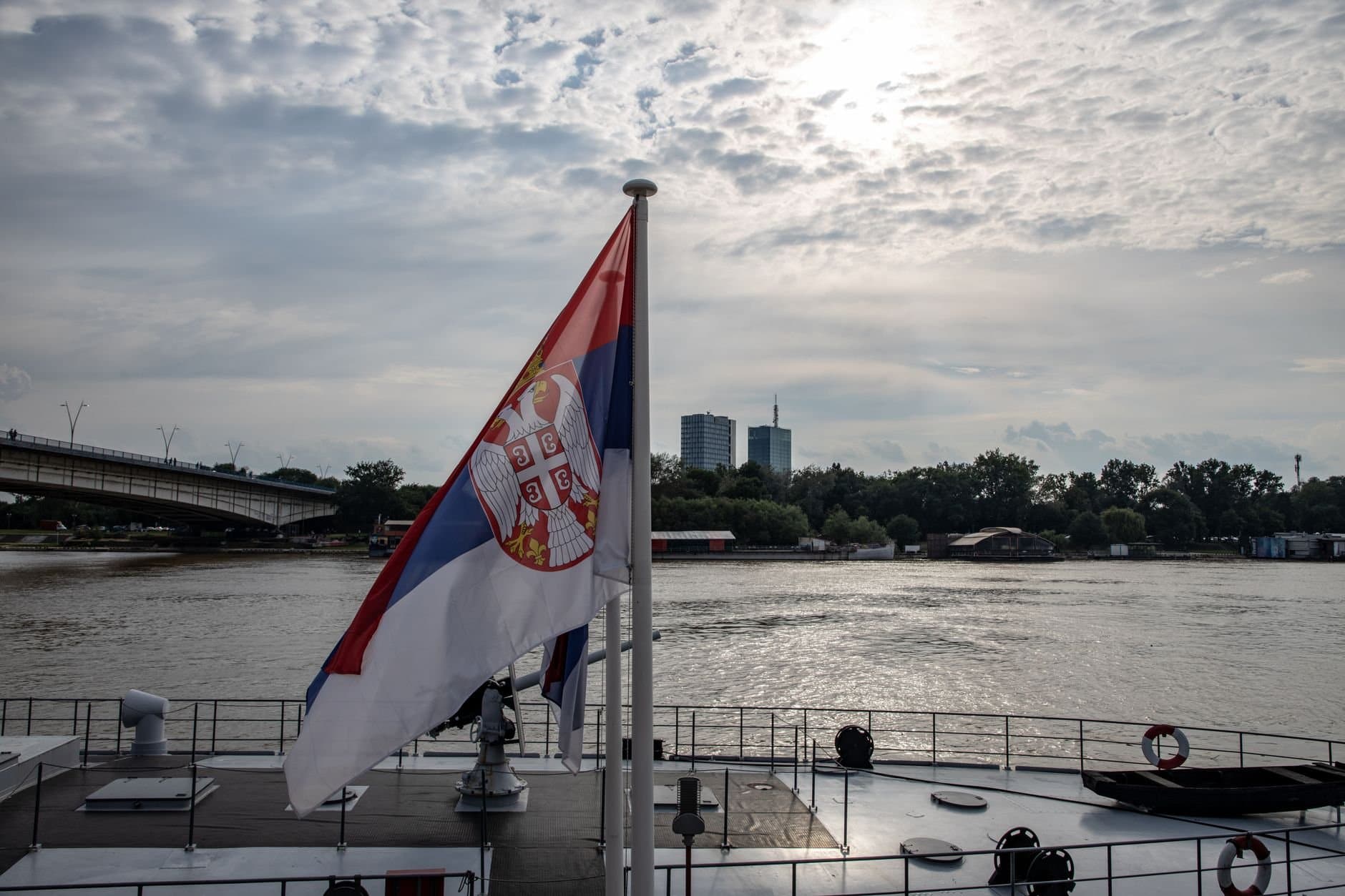 Serbian flag on a boat with an urban skyline and river scene in daytime.