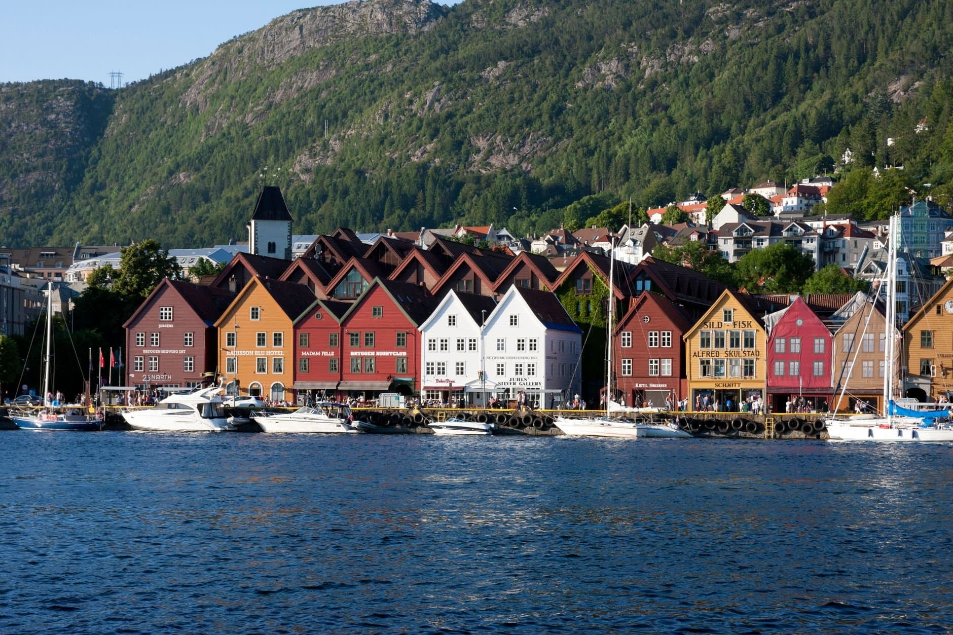 Colorful buildings along Bergen's historic waterfront with boats and lush mountains in view.