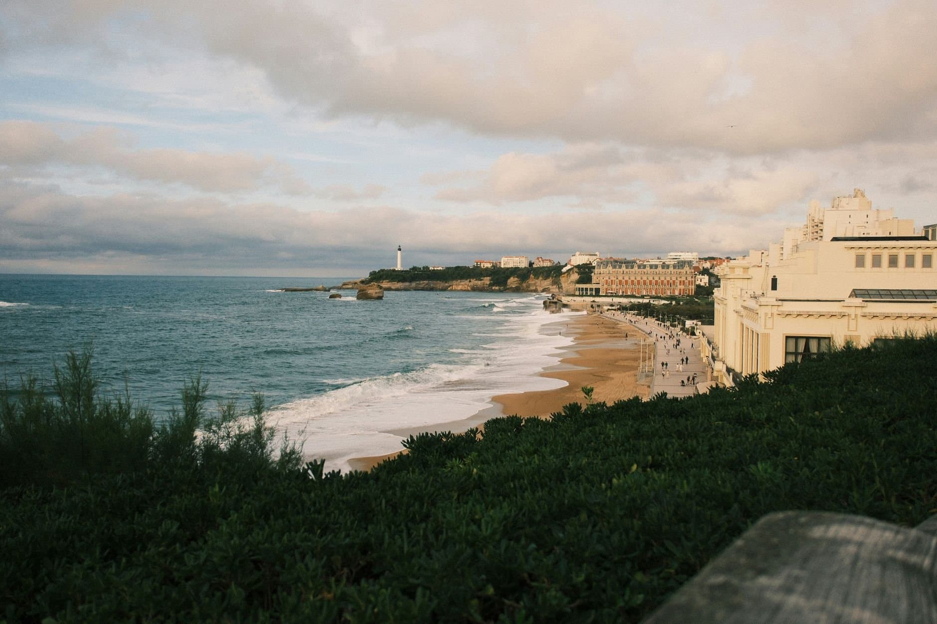 Explore Biarritz's scenic coastal view with sandy beaches and lighthouse under a cloudy sky.