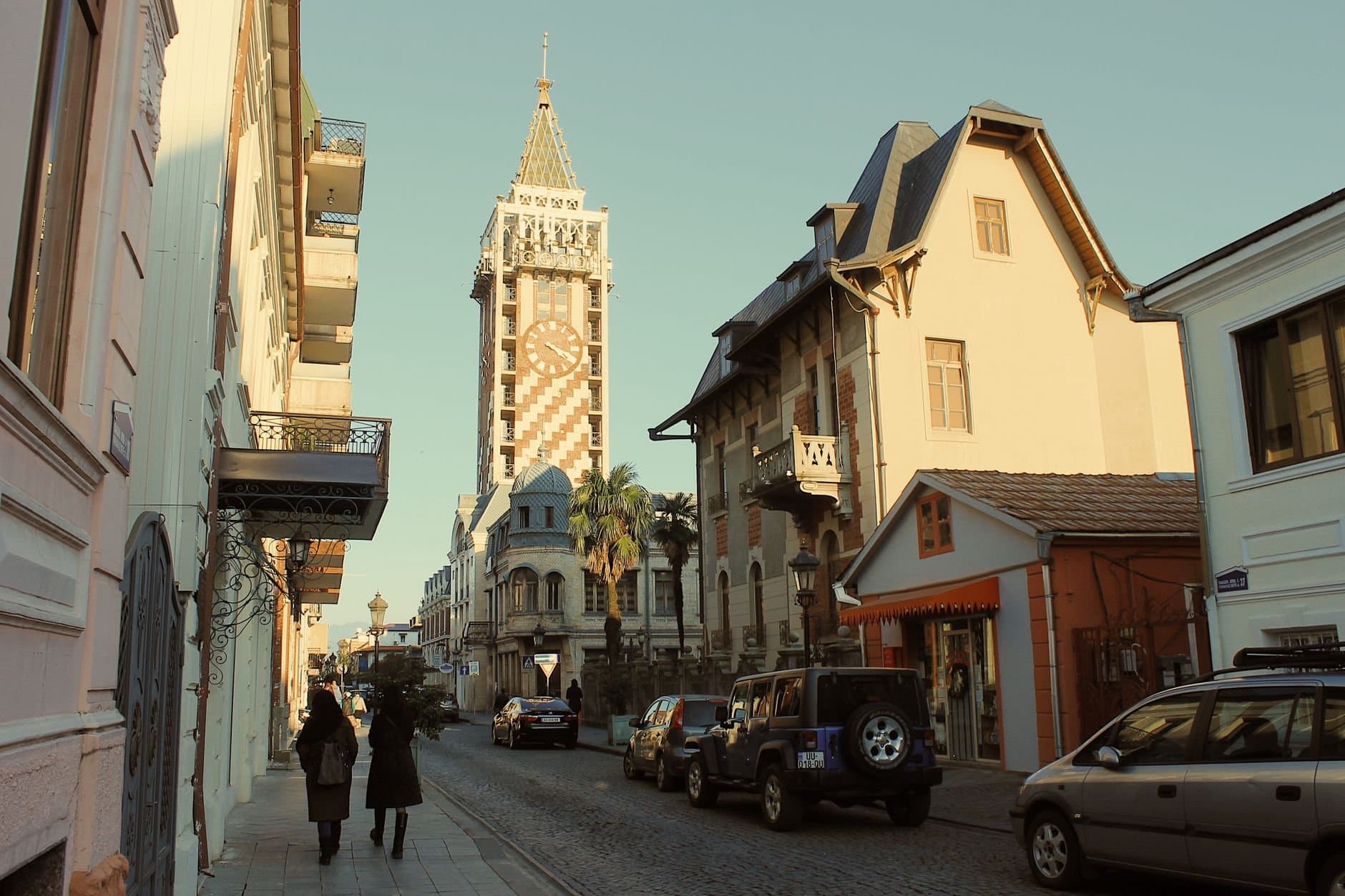 A picturesque street scene featuring a clock tower and historical buildings in a warm, golden hour setting.
