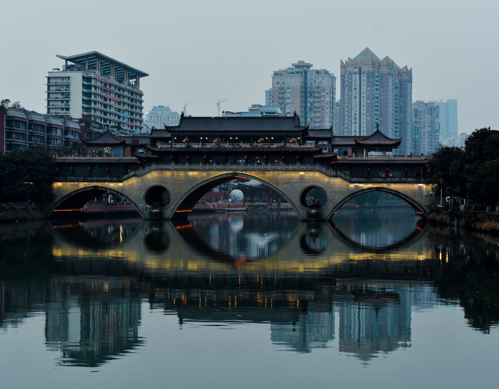 Traditional bridge and modern city skyline reflecting in a serene river in Chengdu.