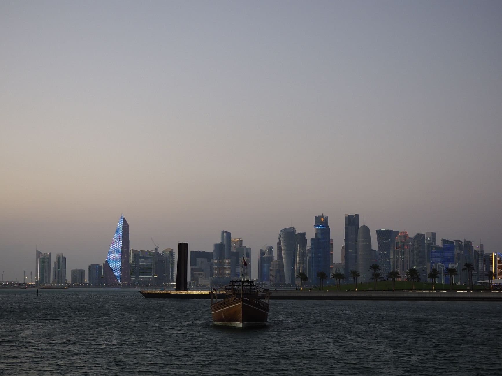 Scenic view of Doha city skyline with traditional dhow boat at dusk along the waterfront.