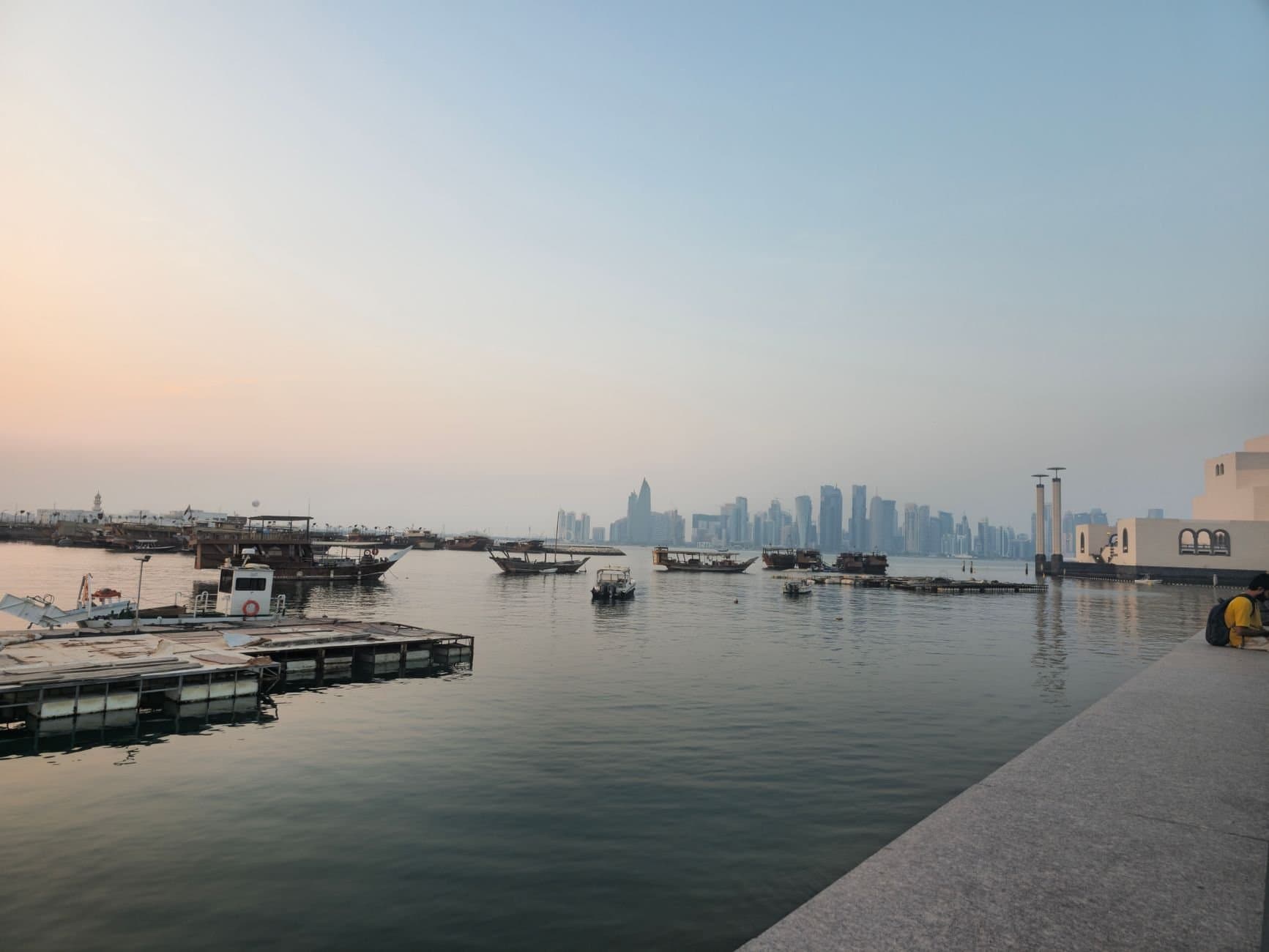 Scenic sunset view of Doha skyline from Corniche with traditional boats and calm water.