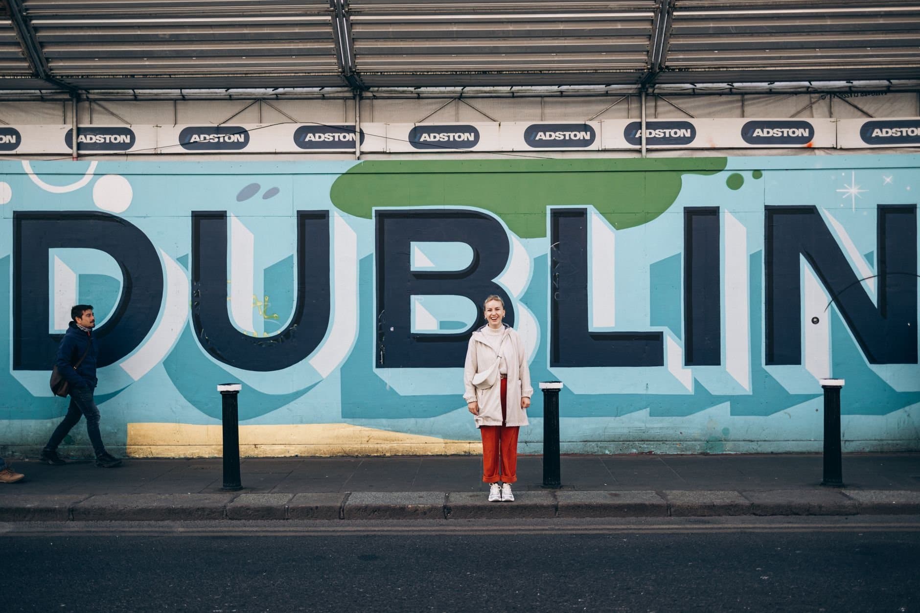 Woman poses in front of colorful Dublin wall mural with passersby on city street.