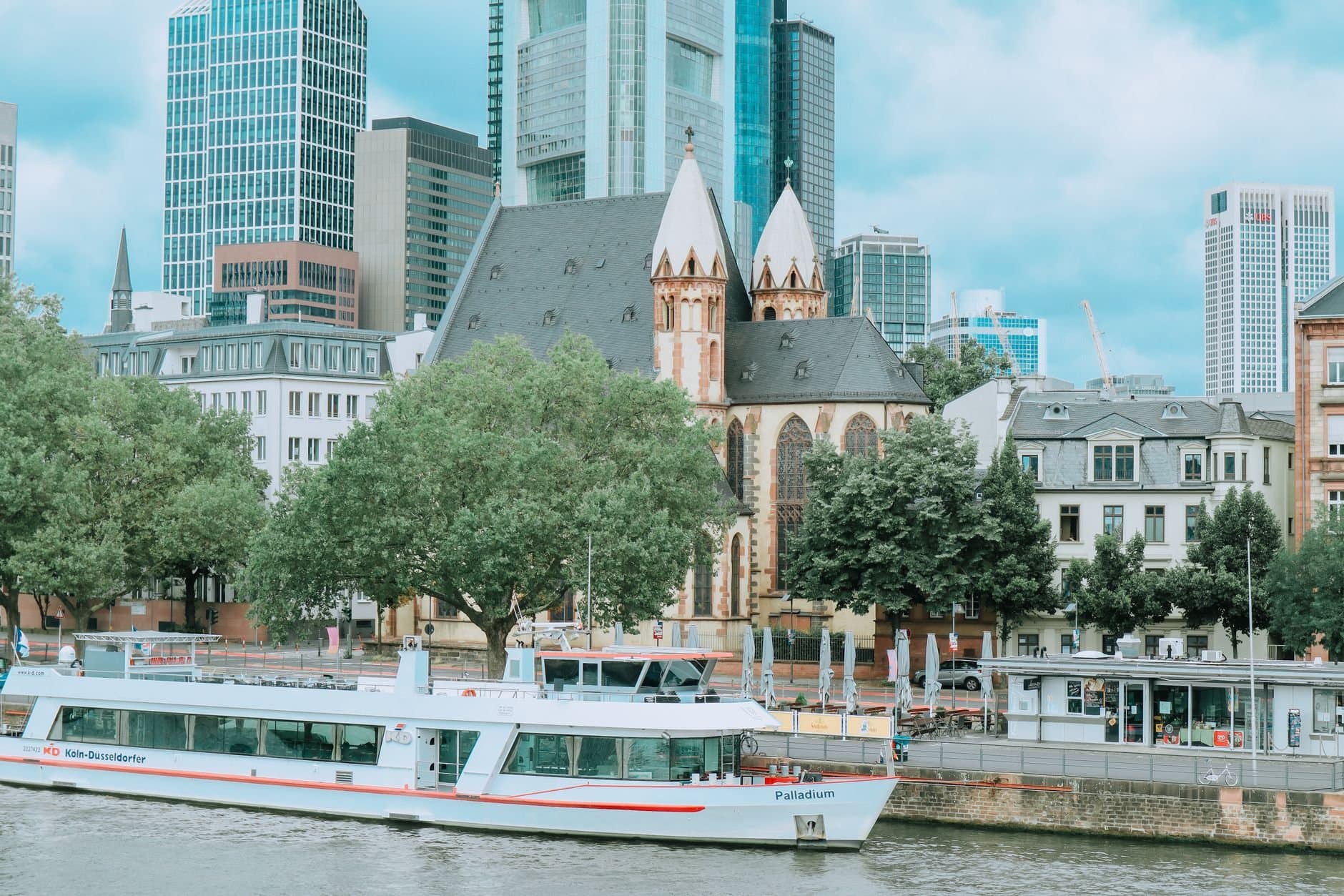 Frankfurt cityscape featuring a historic church, modern skyscrapers, and a boat along the river.