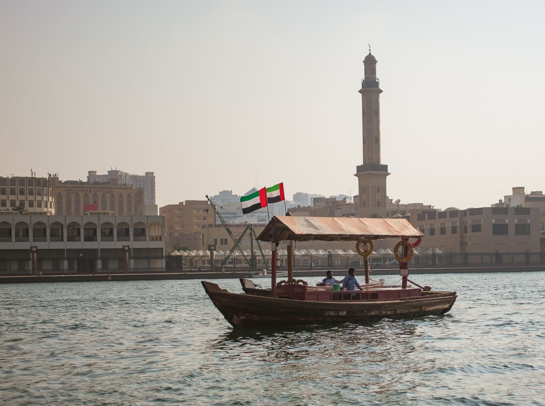 A traditional abra boat sails on Dubai Creek with a city skyline and minaret in the background.