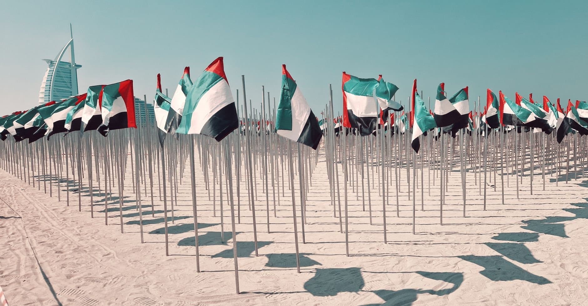 Rows of UAE flags on a sunny beach in Dubai, with Burj Al Arab in the background.
