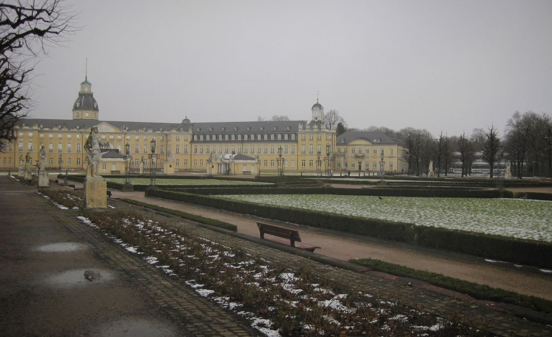 A scenic view of Karlsruhe Palace and garden in winter, showcasing classic architecture under a cloudy sky.