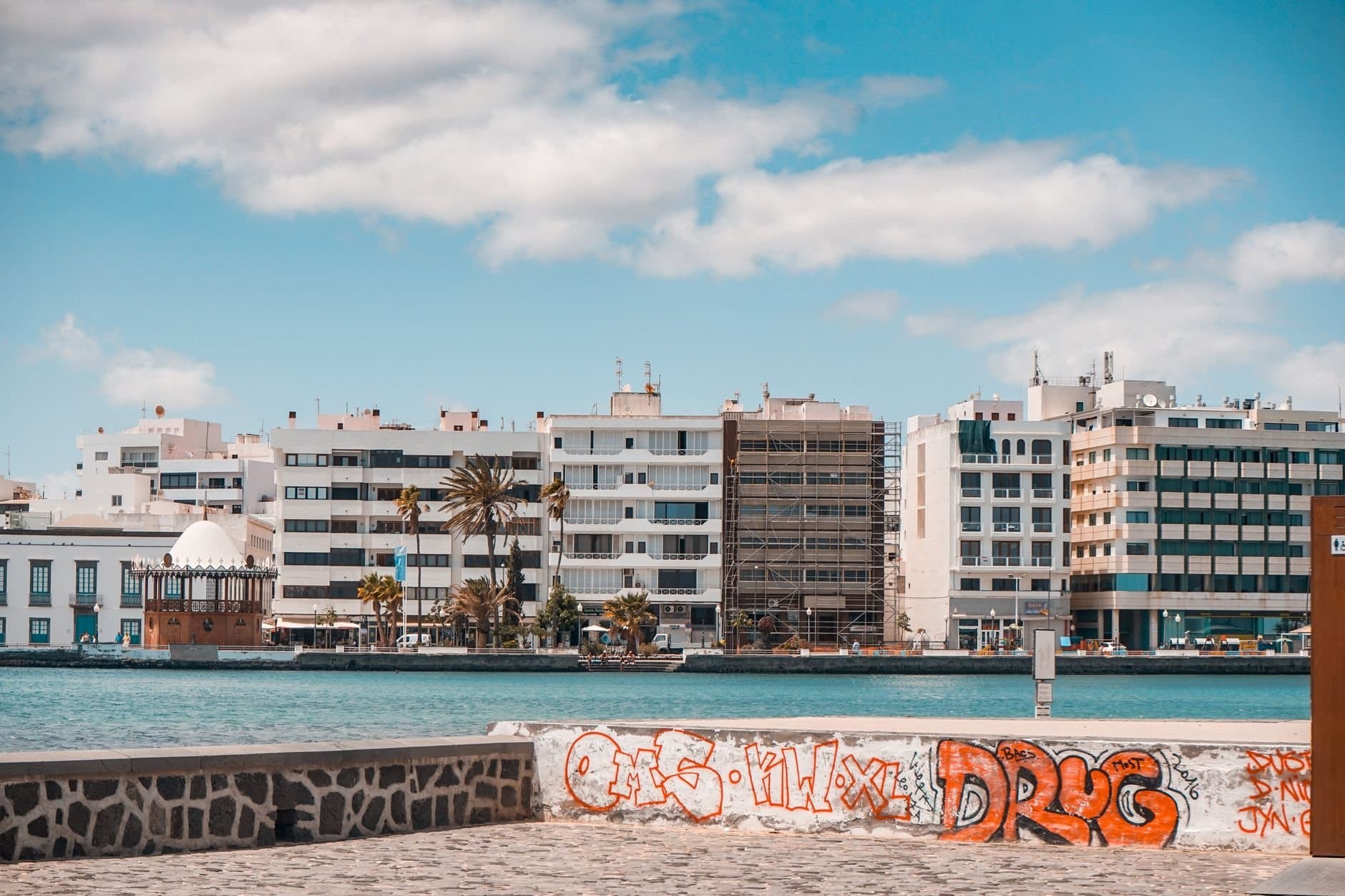 Scenic waterfront of Puerto del Carmen, Spain with buildings and graffiti under a blue sky.