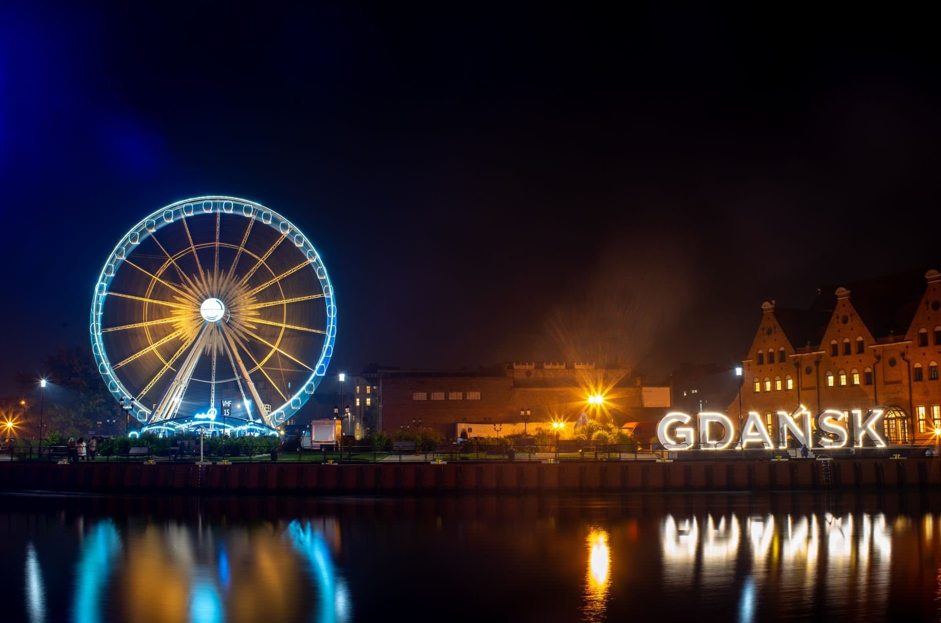 Night view of the brightly lit Ferris wheel and waterfront in Gdansk, Poland, reflecting in the water.