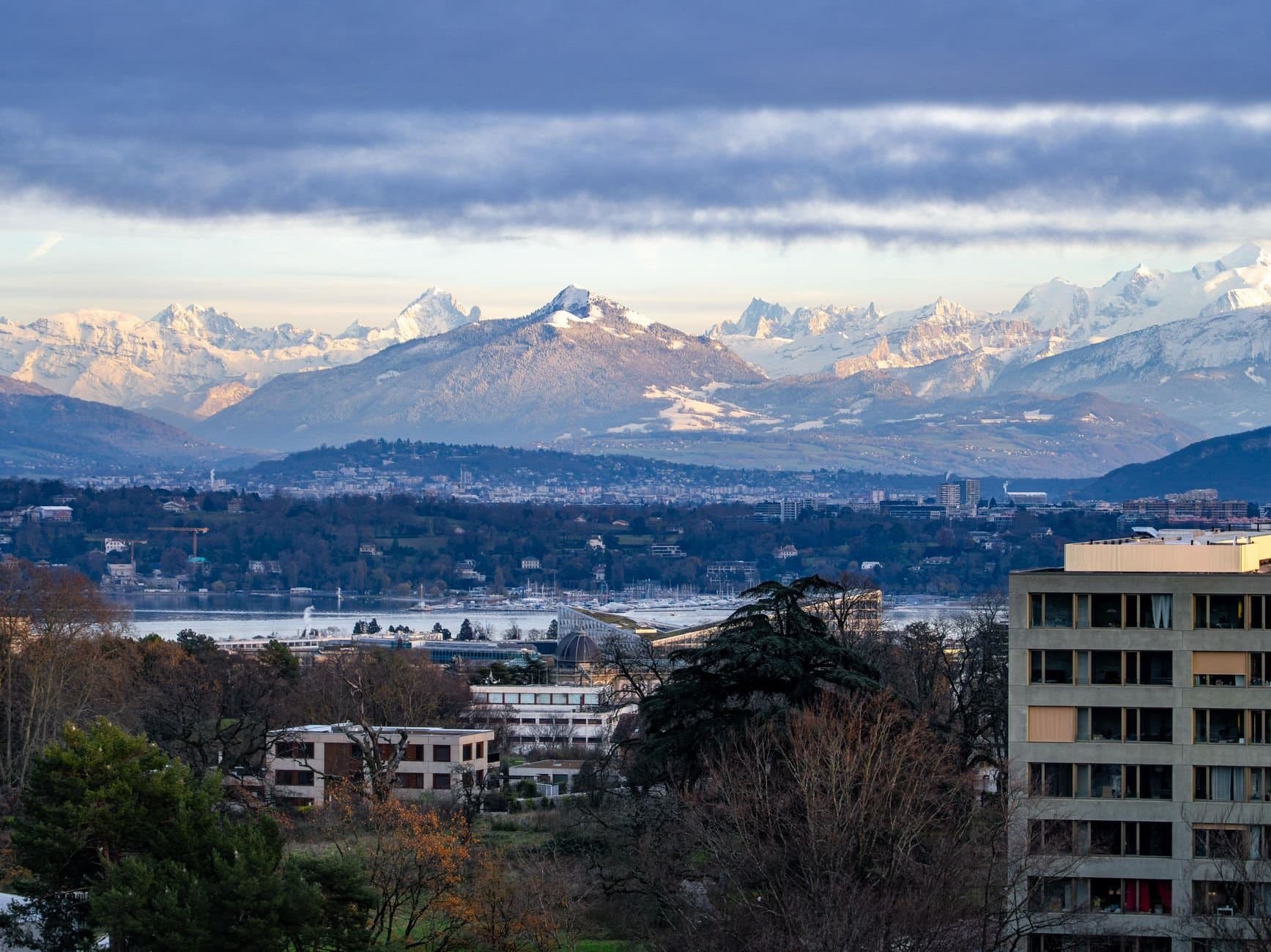 A breathtaking view of Geneva with Swiss Alps backdrop under a cloudy sky at day.