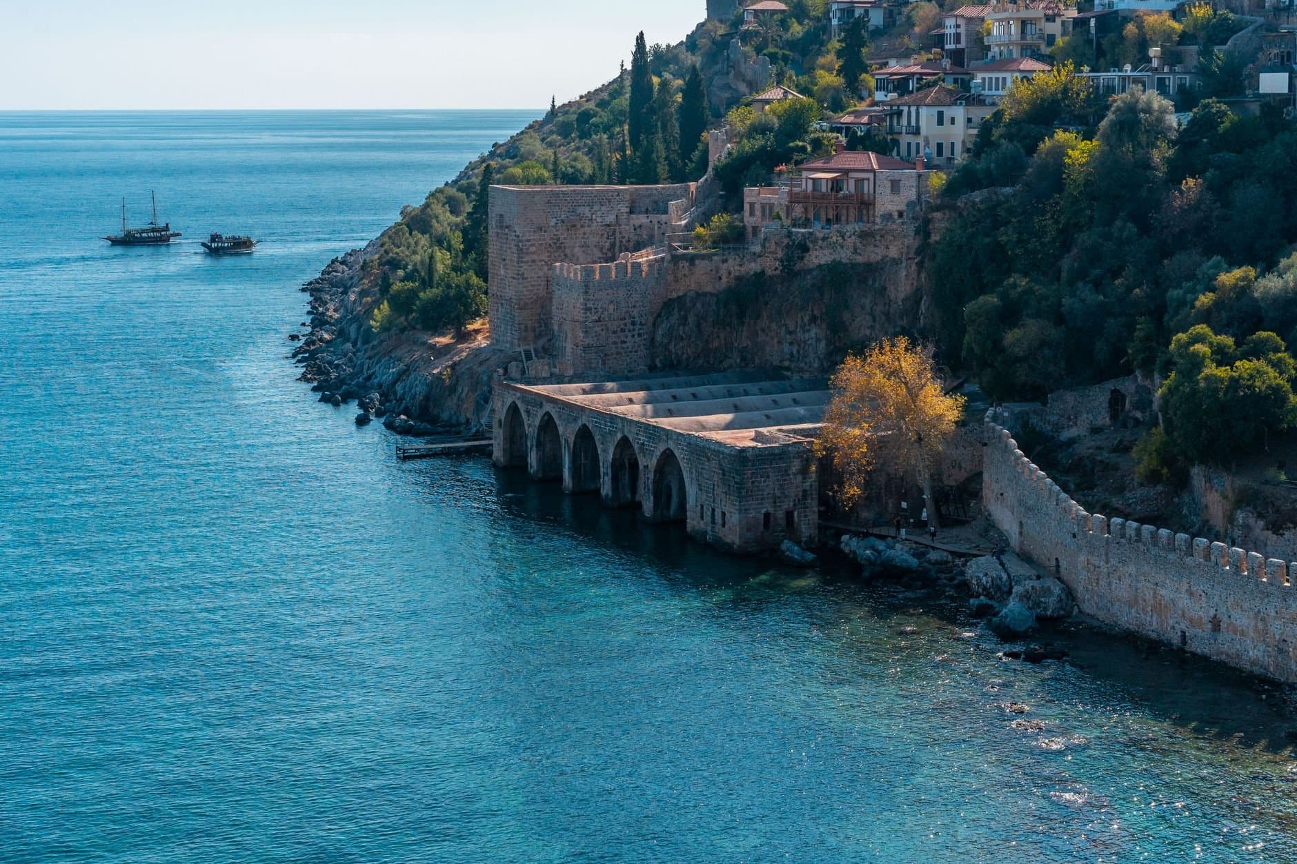 Weathered stone fortress located on rocky seashore with green trees and small settlement with residential buildings near calm sea with boats