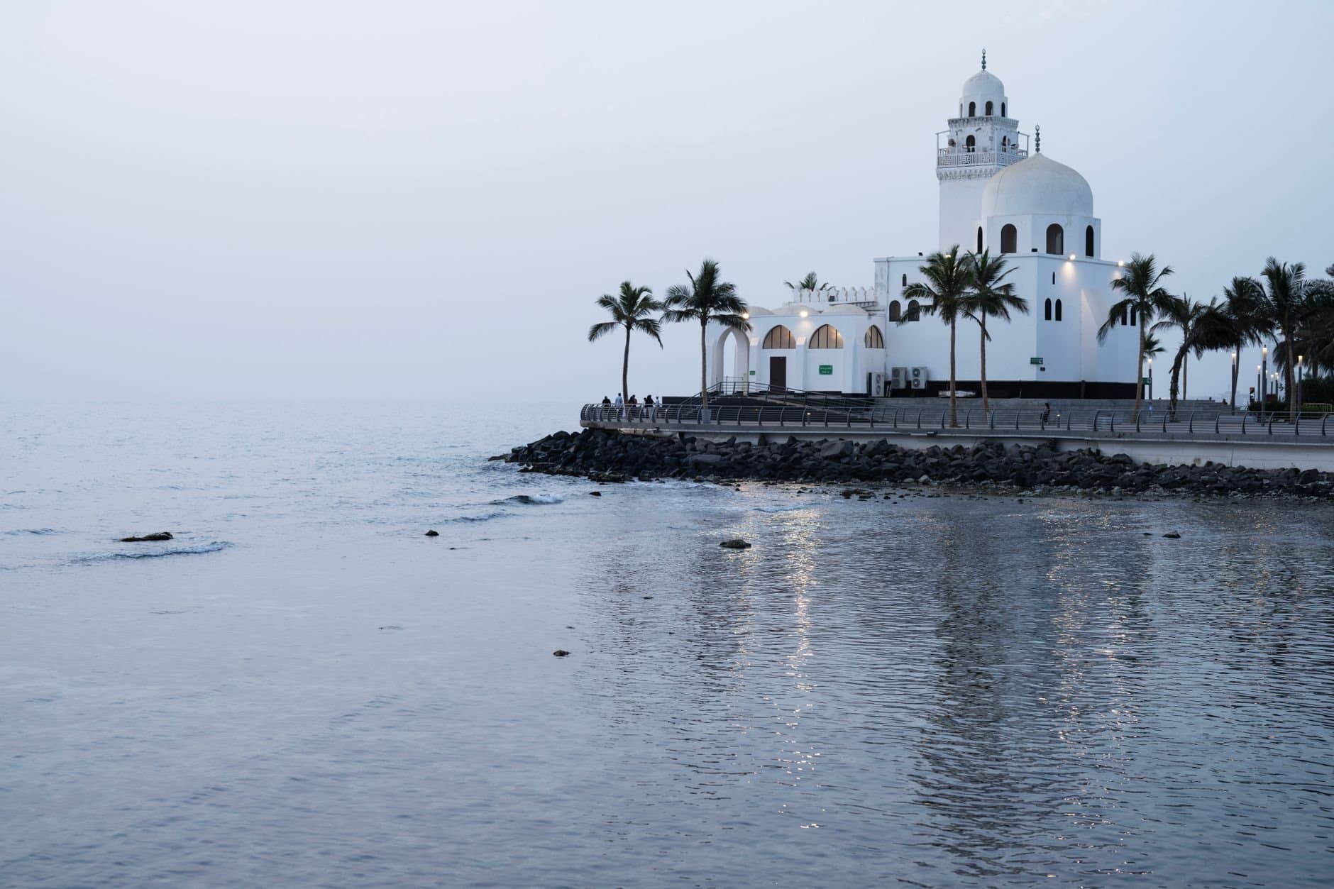 Scenic view of a seaside mosque in Jeddah, Saudi Arabia with palm trees and calm waters.