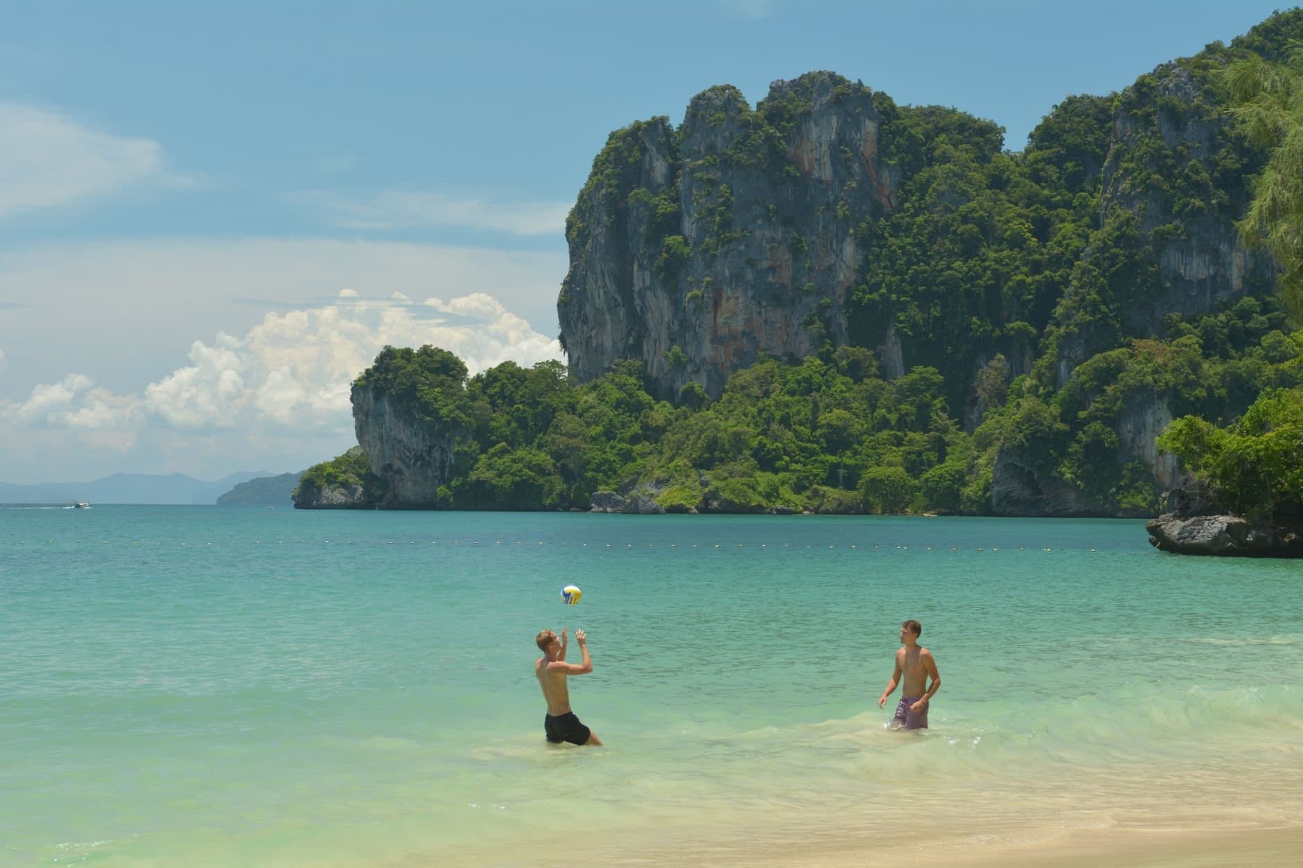 Two men enjoy a game of beach volleyball on Ao Nang's sandy shores with stunning limestone cliffs in the background.