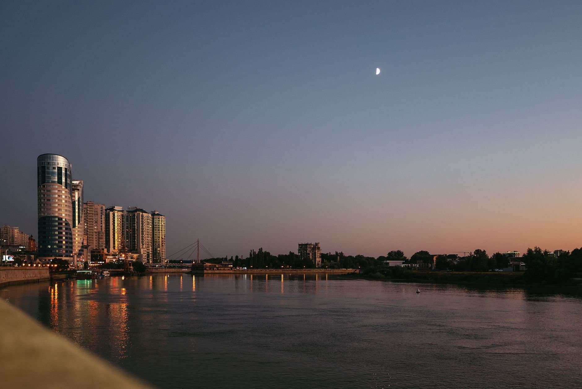 Twilight view of Krasnodar's skyline with reflection on the river and moonlit sky.
