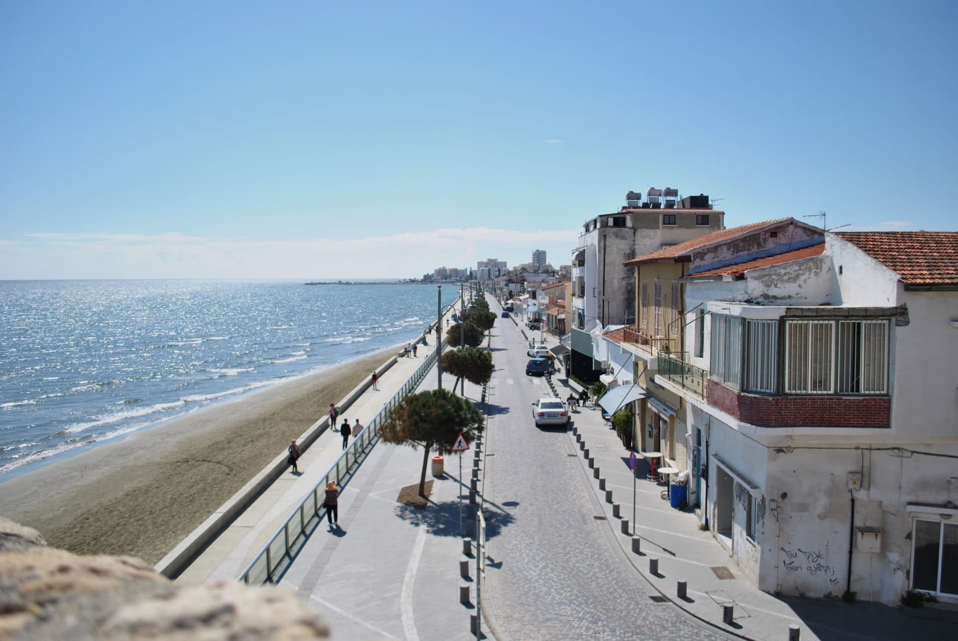 A scenic street along the coast in Larnaca, Cyprus with clear blue skies, waterfront, and walking pedestrians.