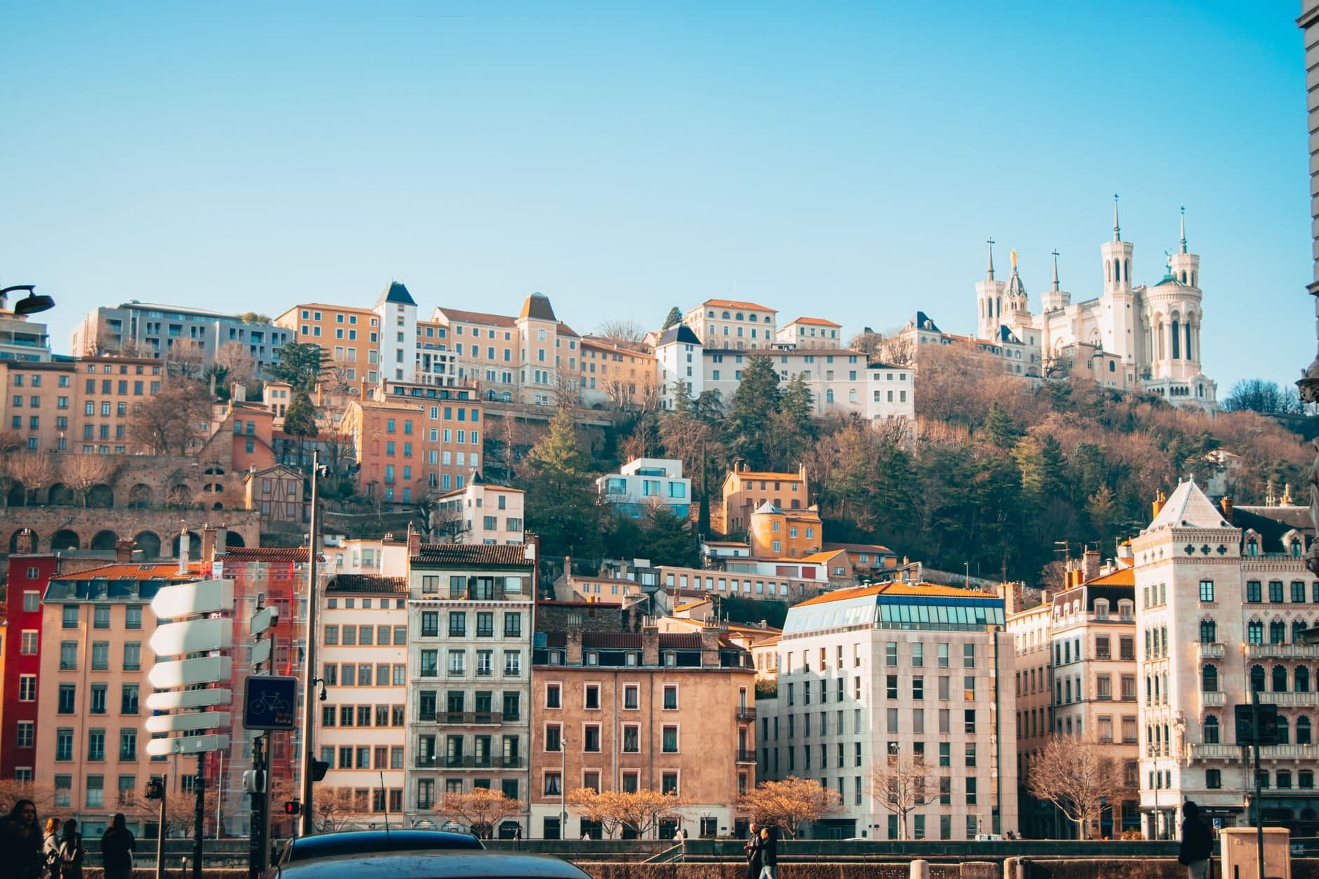 A vibrant cityscape of Lyon featuring the Basilica, buildings, and lush greenery on a hill.