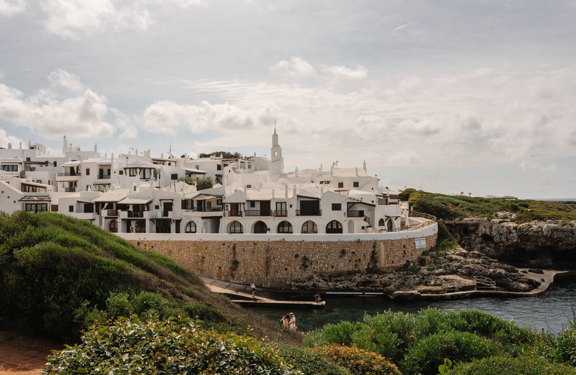 Scenic view of white buildings along the coast in Ciutadella de Menorca, Spain, with lush greenery and calm waters.