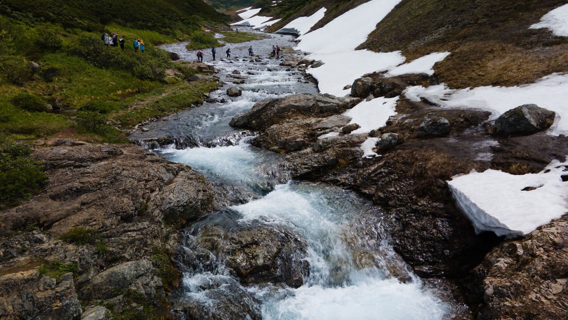 A breathtaking view of a flowing mountain stream surrounded by snow patches and hikers in Kamchatka.