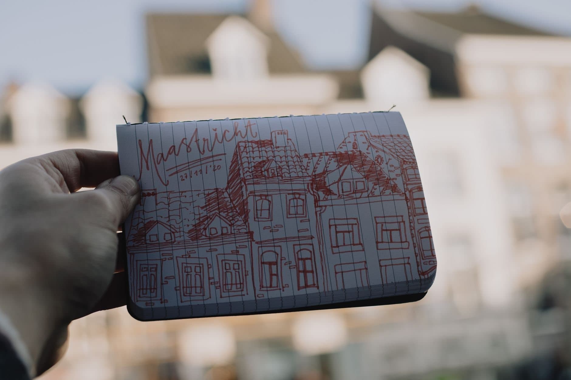 A hand holds a sketch of Maastricht architecture outdoors with blurred background.