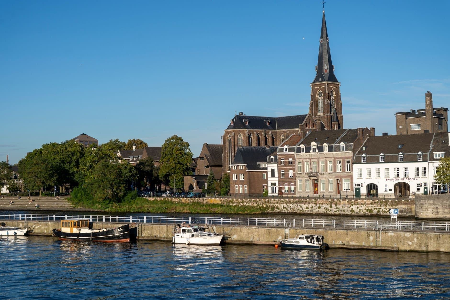 Discover Maastricht's charming riverside with historic buildings and the iconic church under a bright summer sky.