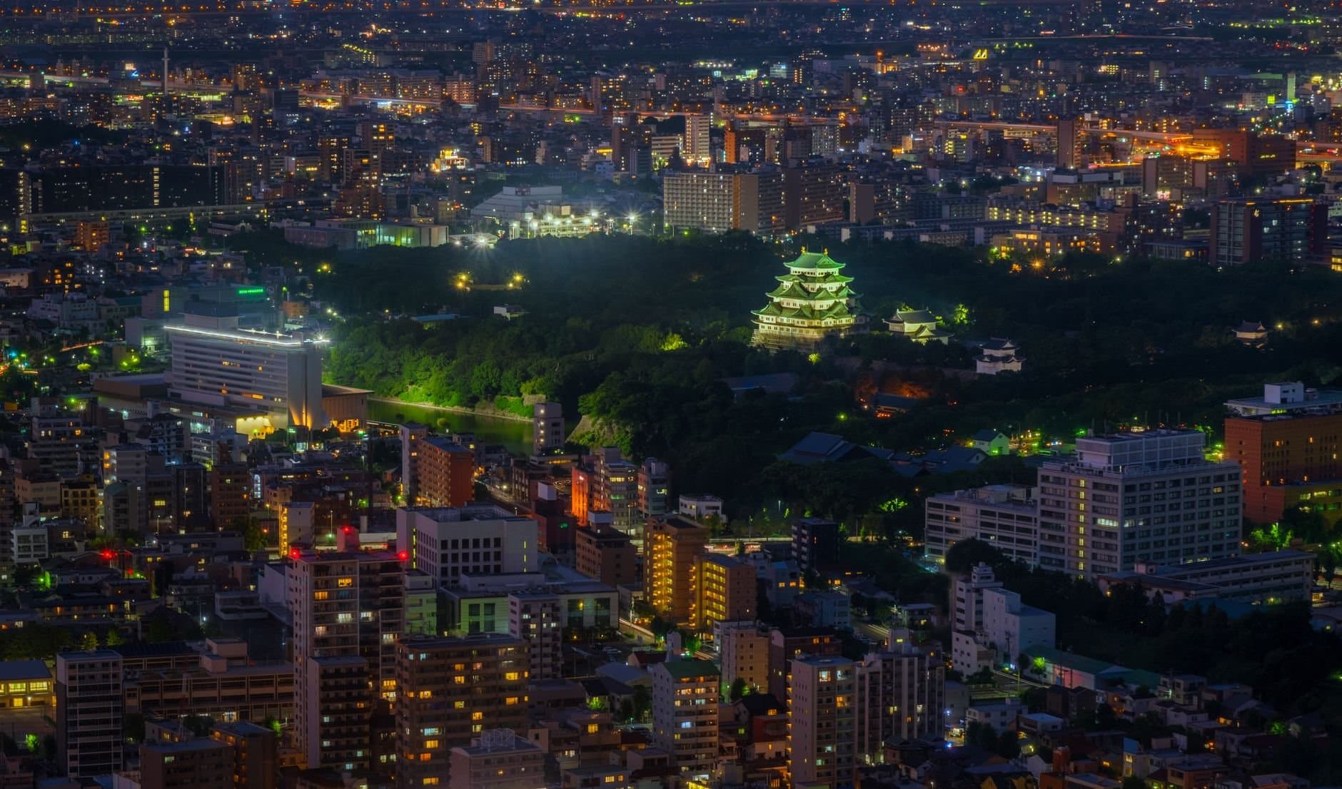 Aerial shot of illuminated Nagoya Castle amidst urban cityscape at night.