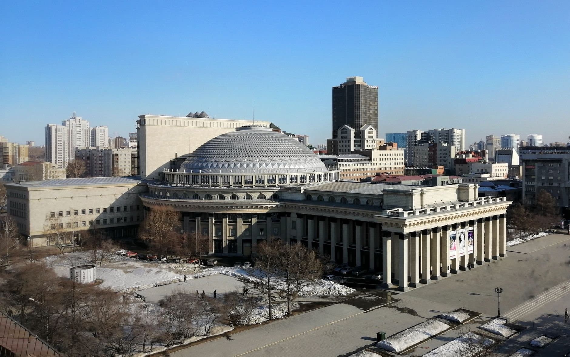 Aerial view of the Novosibirsk Opera House, showcasing its dome and urban skyline under a clear winter sky.