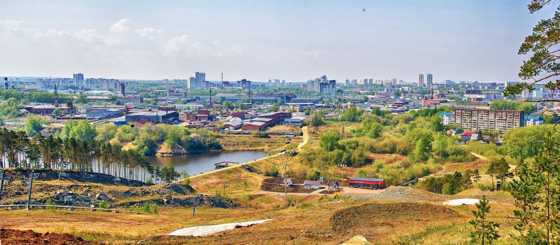 Panoramic aerial view of a cityscape with a lake, surrounded by lush greenery and urban architecture.