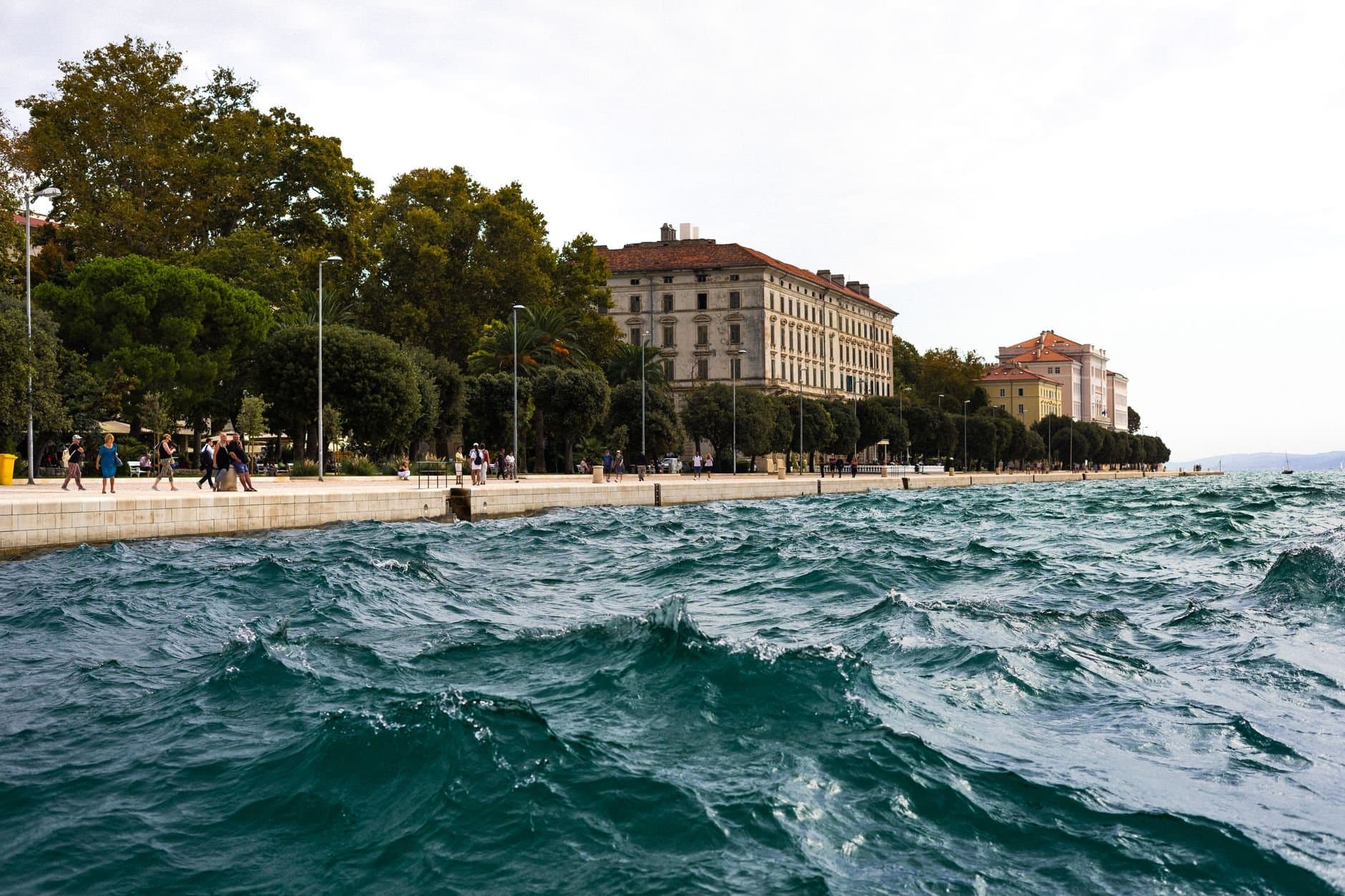 Waves crashing against a scenic coastal promenade lined with trees and historic buildings.