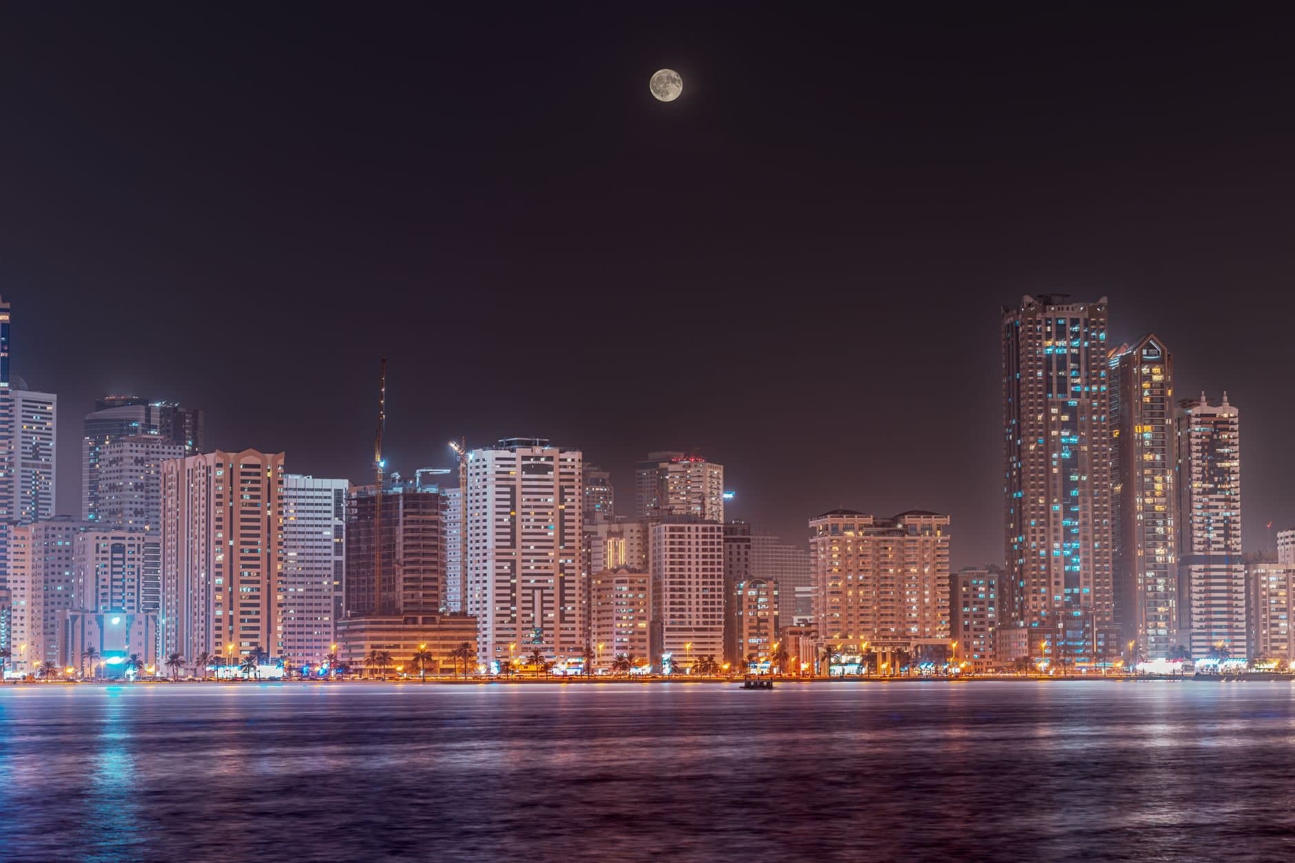 Scenic night view of Sharjah skyline with a full moon reflecting on the water.