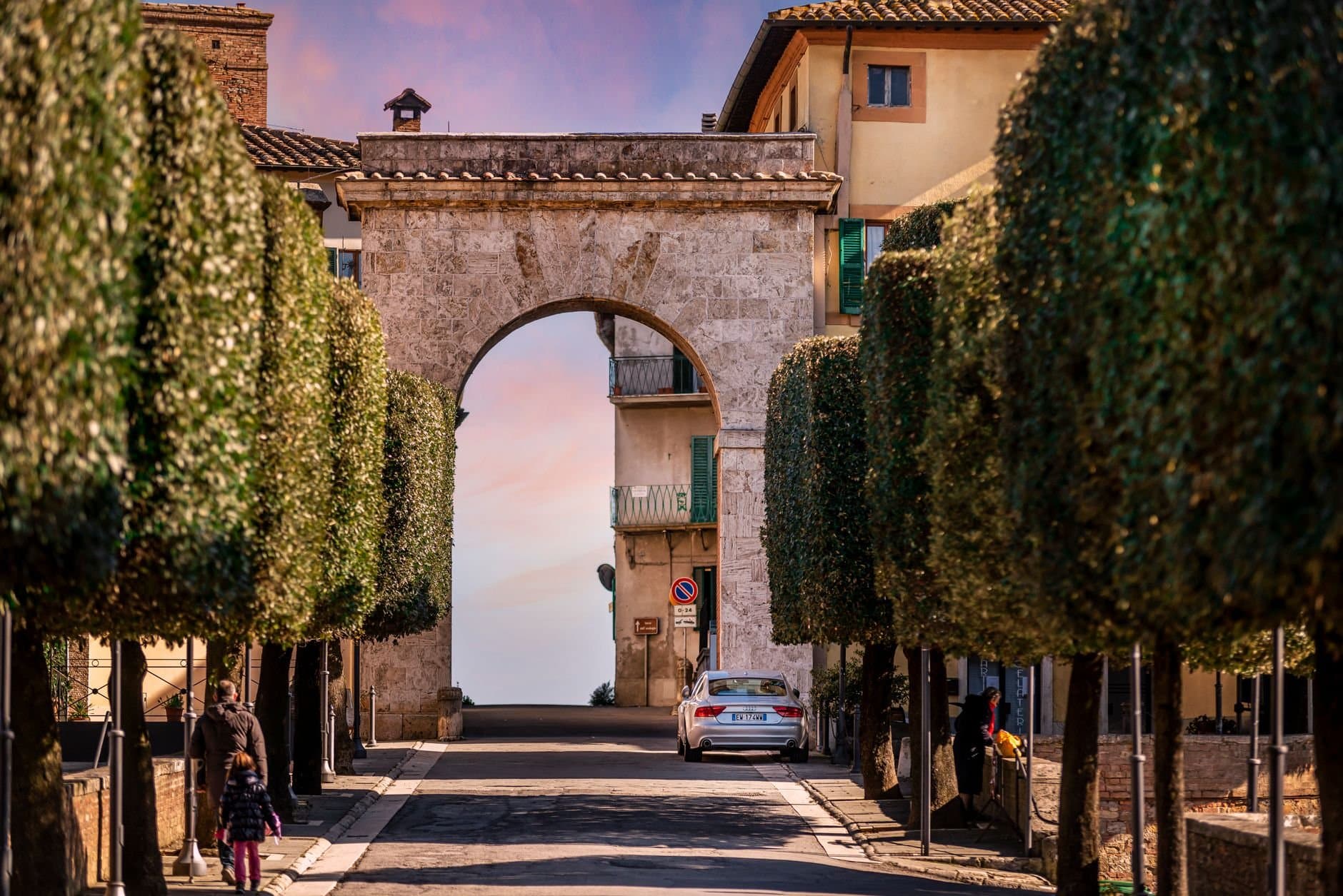 Narrow cobblestone street amidst lush green trees leading to stone arch against sunset sky in old town of Chianciano Terme