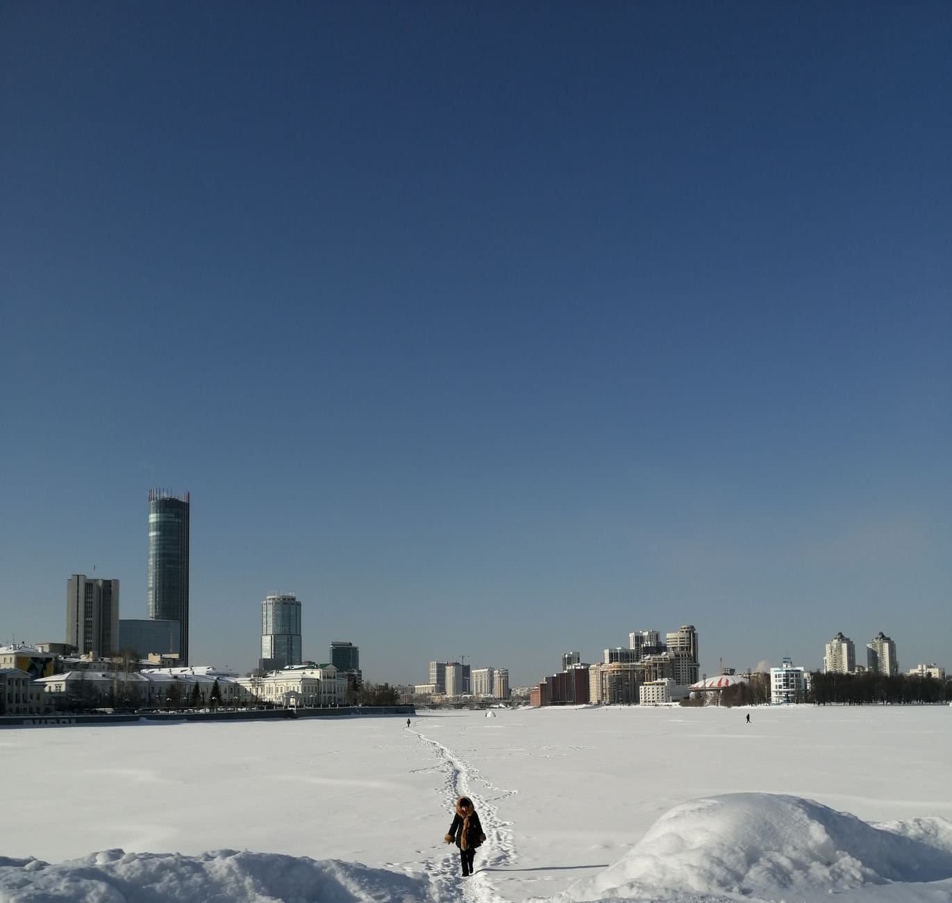 Woman walking in snow-covered field near Yekaterinburg skyline under a clear winter sky.