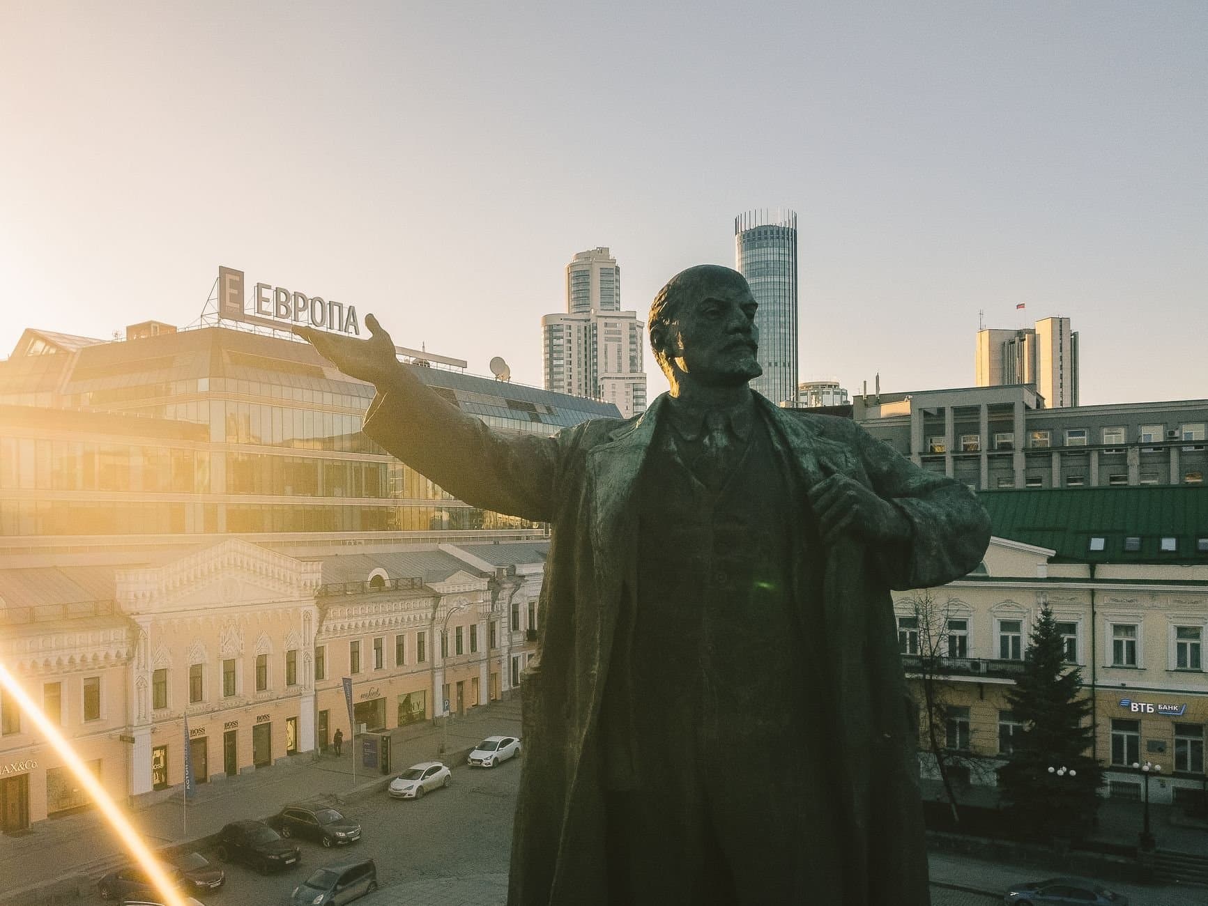 A Lenin statue overlooks Yekaterinburg cityscape under golden daylight, highlighting the historic and urban blend.