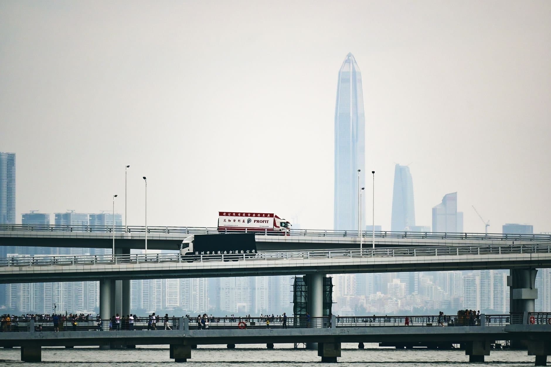 A truck crosses a double-decker bridge against a modern city skyline with high-rise buildings.