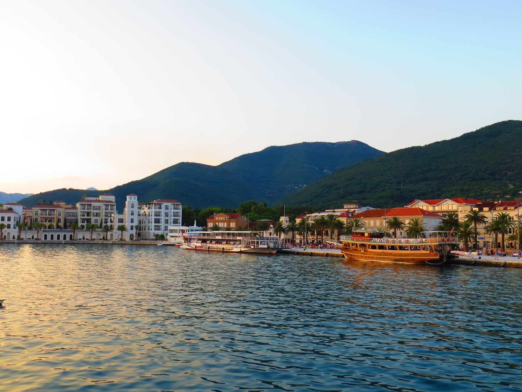 Picturesque view of modern coastal town surrounded by green hills and located near rippling sea in evening sun