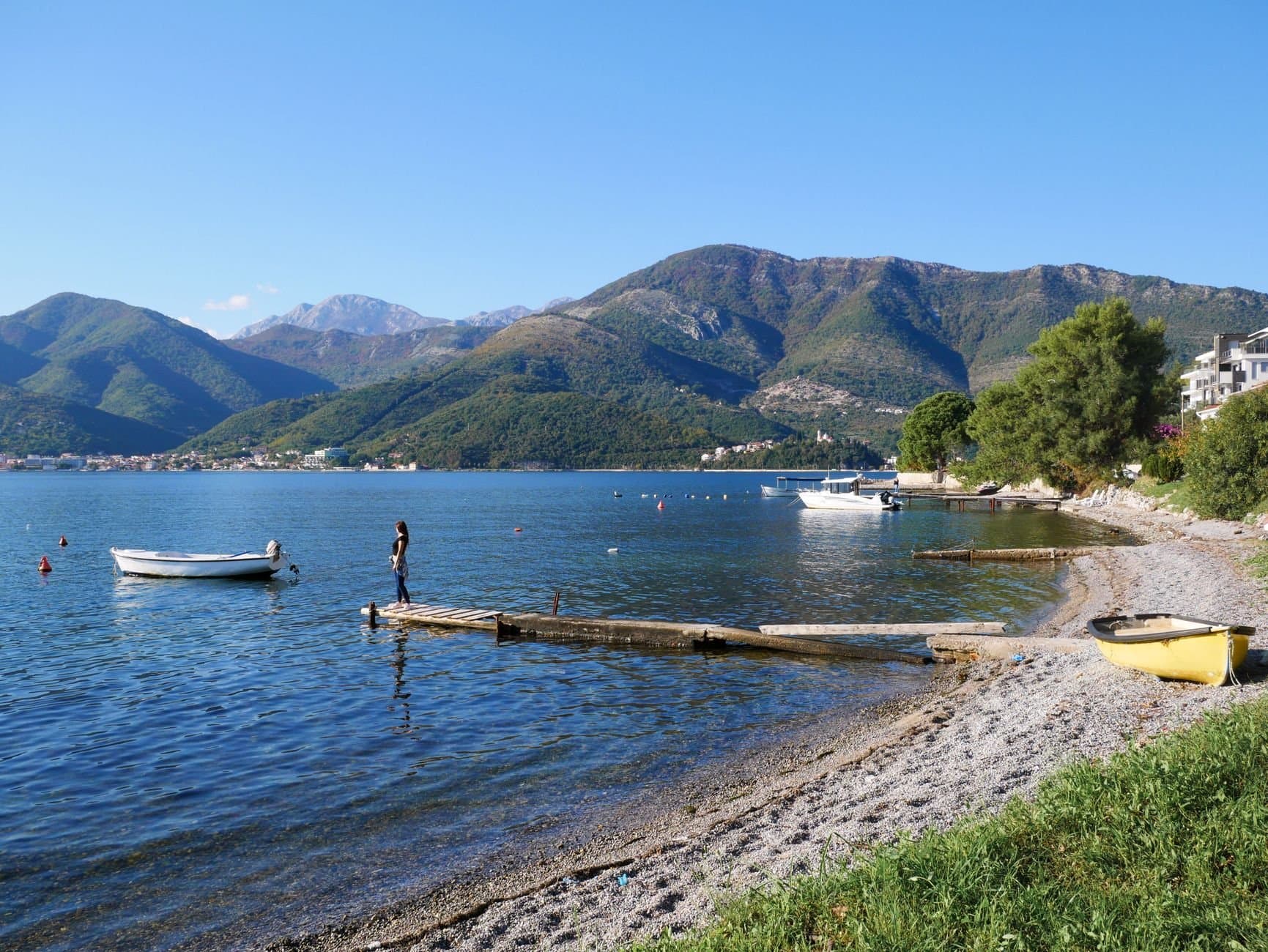 A tranquil beach scene in Tivat with boats and mountains in the background under a clear sky.