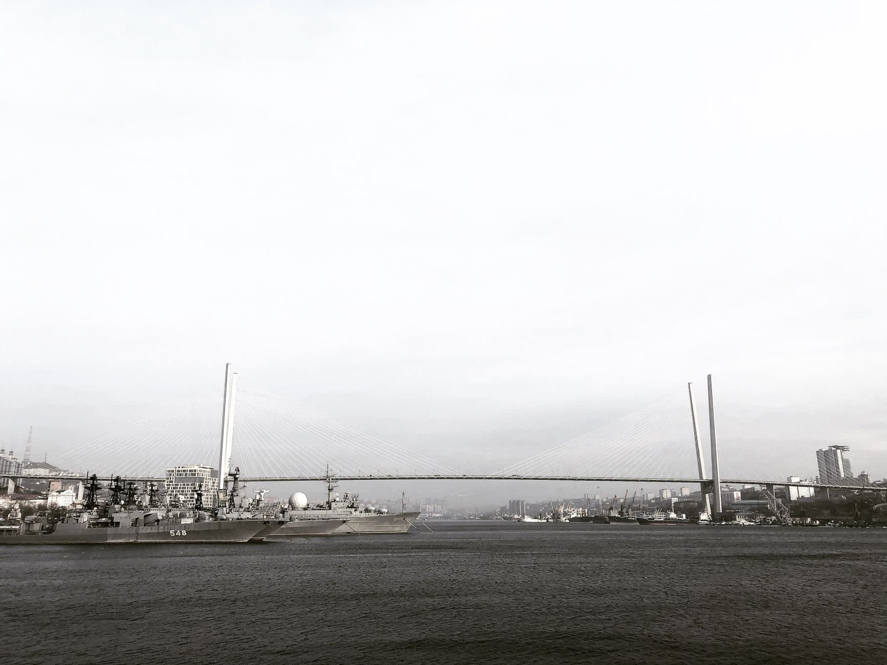 Black and white photo of Vladivostok harbor featuring docked ships and a suspension bridge.