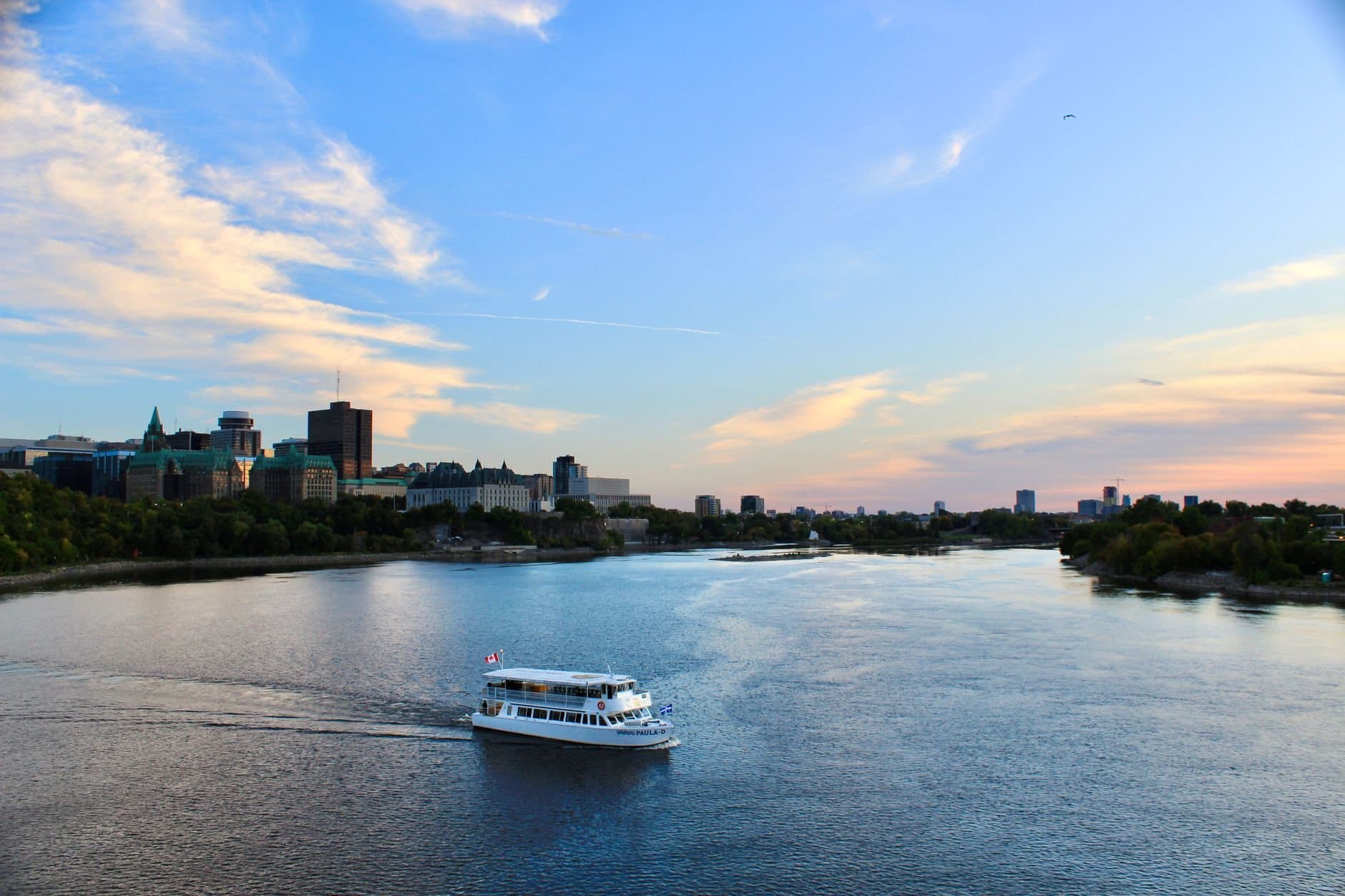 A touring boat cruises on a river with a city skyline backdrop under a bright sky.